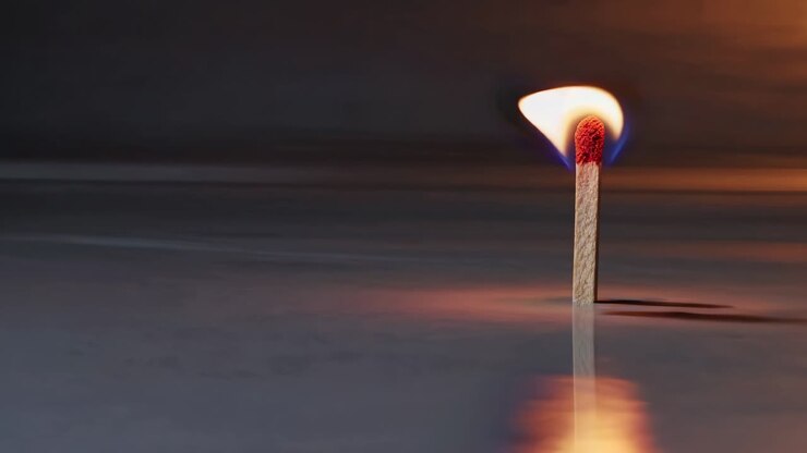 Close-up, low-angle shot of a single lit match on a reflective surface, creating a dramatic
