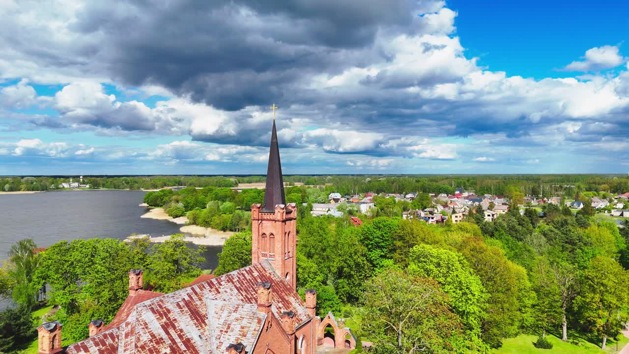 A gothic red brick church with a striking black steeple stands beside a green park under dramatic spring clouds and bright blue skies in a European landscape.