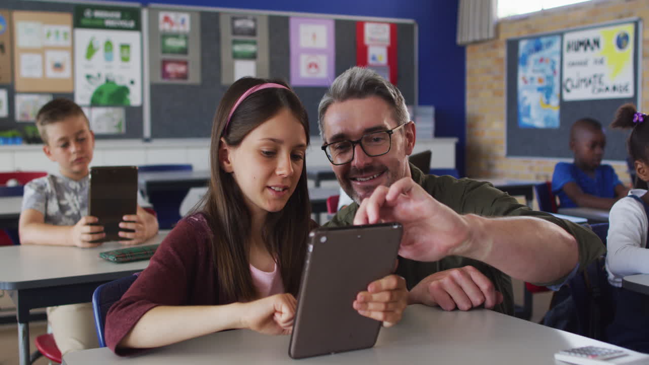 un maestro masculino diverso ayudando a una escolar sentada en el aula usando una tableta