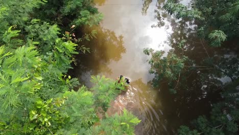 Lost-woman-with-female-child-walking-along-overflowed-road-surrounded-by-forest-trees-during-sunlight-after-rain