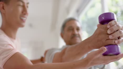 Diverso-Fisioterapeuta-Masculino-Asesorando-Y-Determinado-Hombre-Mayor-Usando-Pesas,-En-Cámara-Lenta