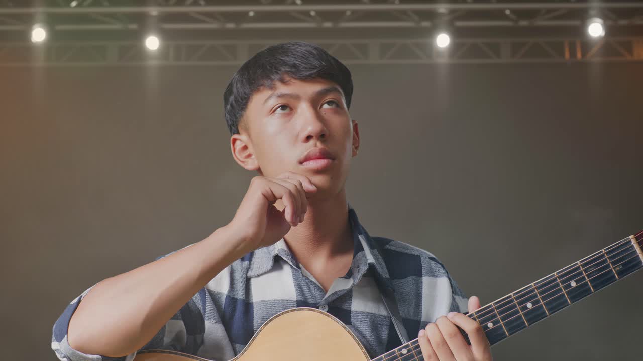 기타를 들고 있는 아시아 십대가 무대에서 무언가를 생각하고 주변을 둘러보고 있다.