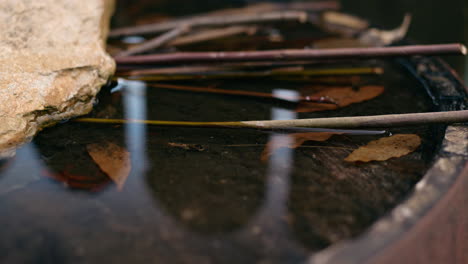 Water-surface-with-a-rock-and-branches-in-a-rusted-metal-barrel,-capturing-reflections-and-natural-light-on-a-calm,-overcast-day-outside