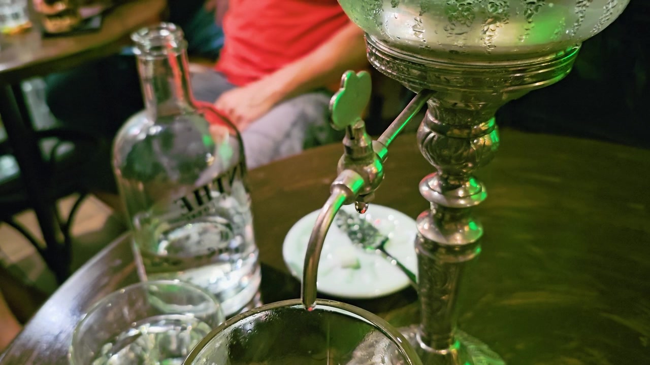 Detaillierte Baraufnahme einer Absinthfontäne, die gekühltes Wasser in ein Cocktailglas gießt, mit verziertem Metallausguss, Flasche, Glaswaren, sanften grünen Reflexionen und gedämpfter Nachtleben-Atmosphäre