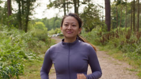 Mujer-De-Mediana-Edad-Haciendo-Ejercicio-Al-Aire-Libre-Corriendo-Por-Una-Pista-A-Través-Del-Bosque-Hacia-La-Cámara-Vistiendo-Ropa-Deportiva