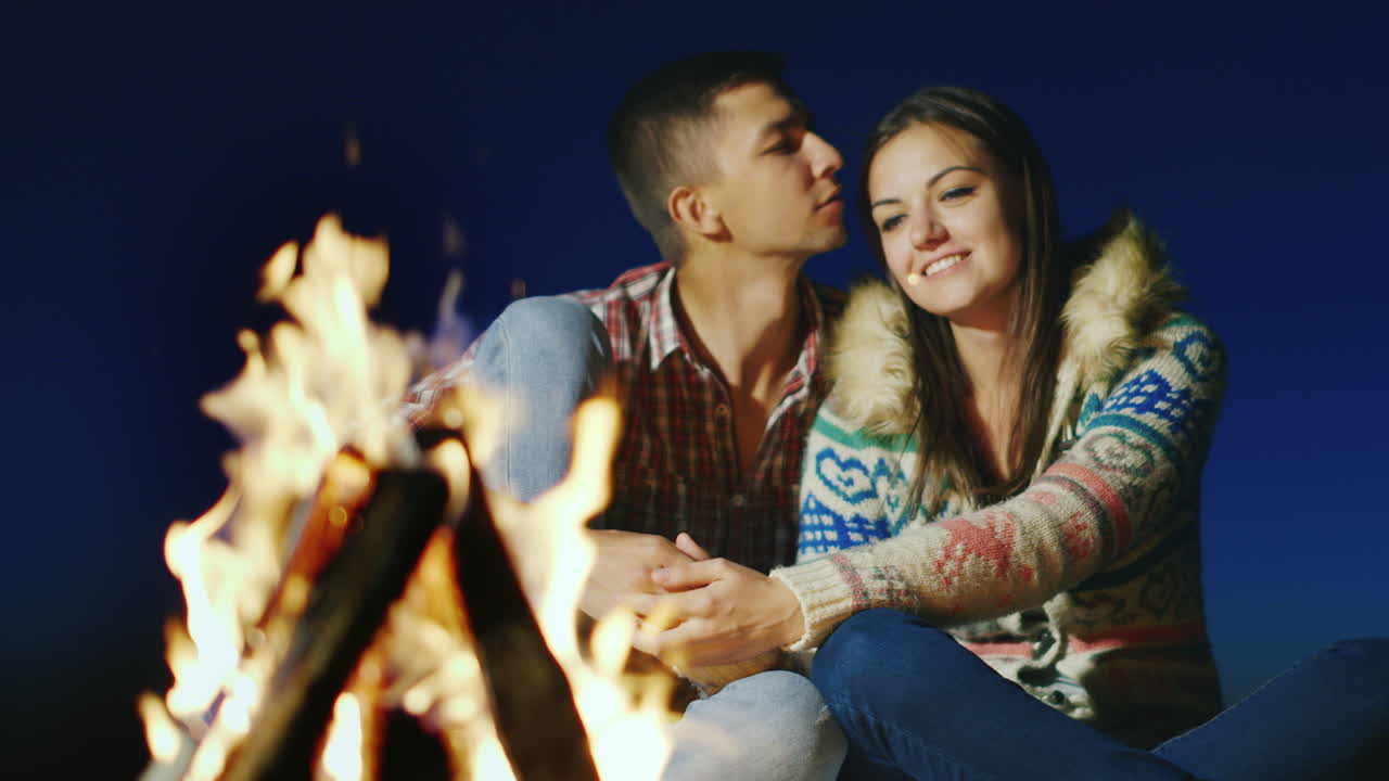 una pareja amorosa sentada junto al fuego tomados de la mano admirando el fuego
