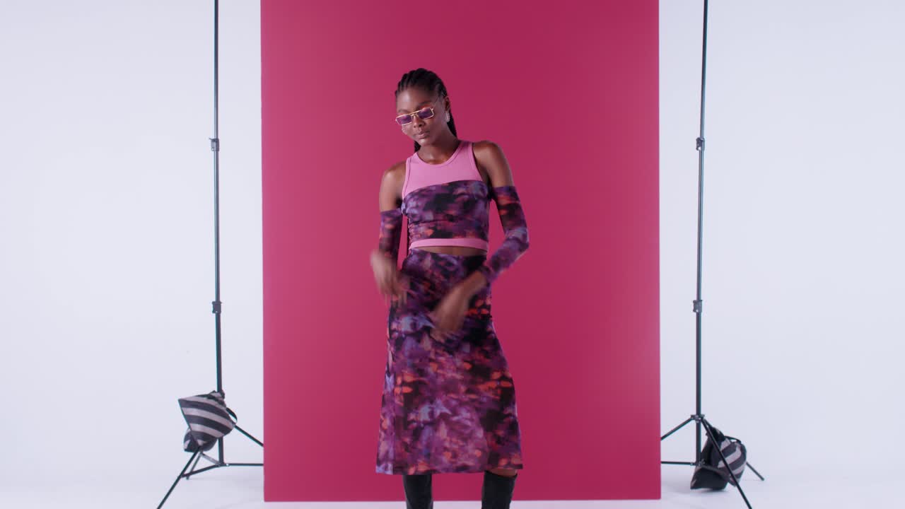 Stylish Black woman poses with sunglasses, smiling brightly and moving her arms against a vibrant pink background