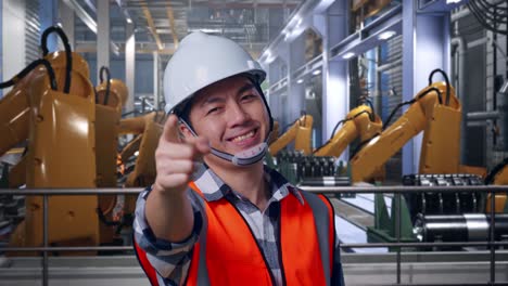 close up of asian male engineer with safety helmet smiling and touching his chest then pointing at you while standing with industrial robotic arms working