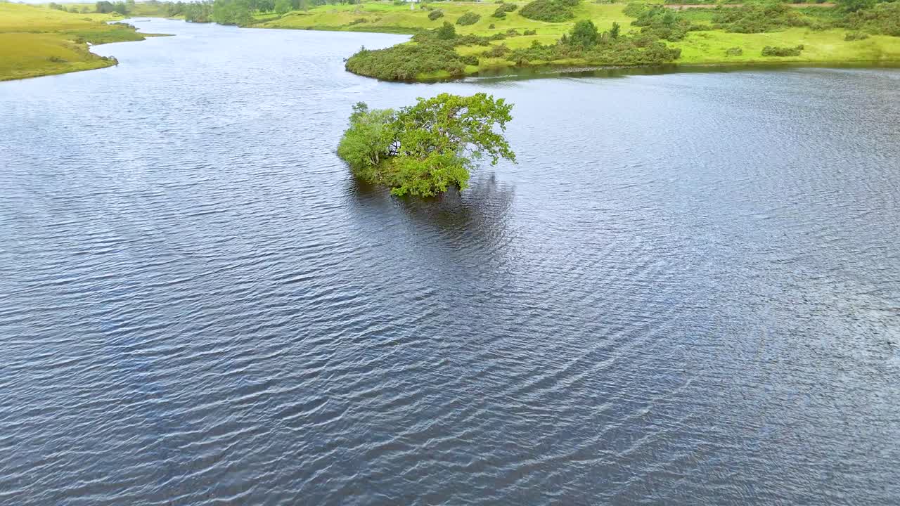 Drone glides over rippling water toward a small tree-covered island in a lush Highland wetland under diffuse daylight, capturing tranquil natural scenery