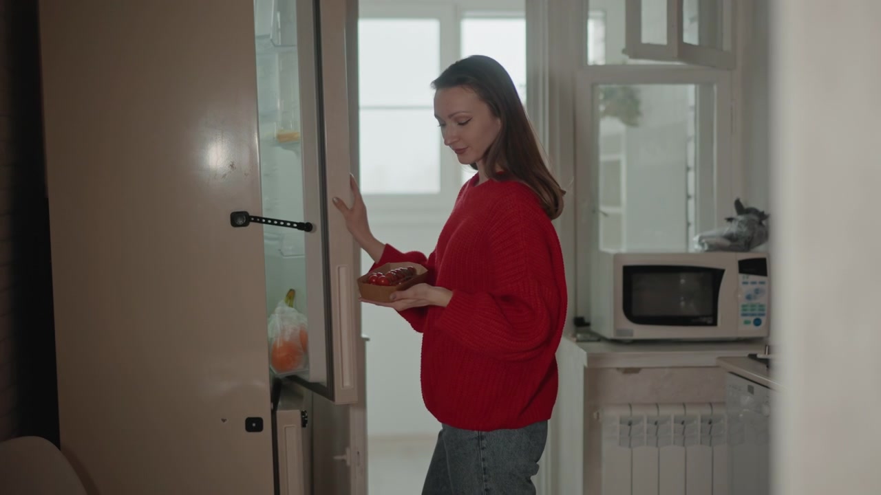 Mladá žena v červeném svetru stojí uvnitř otevřených skleněných dveří v jasném interiéru bytu, drží telefon v jedné ruce a druhou gestikuluje, zatímco se dívá stranou s mírným úsměvem a uvolněným výrazem.