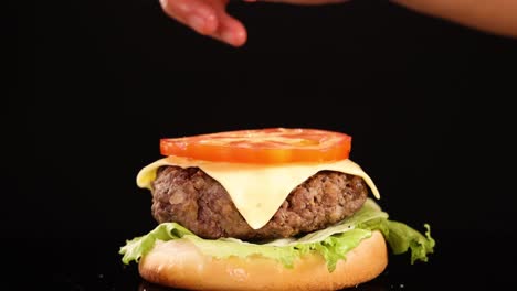 a hand carefully adds a tomato slice onto a cheeseburger with lettuce, cheese, and beef patty, shot in close-up against a black background with studio lighting
