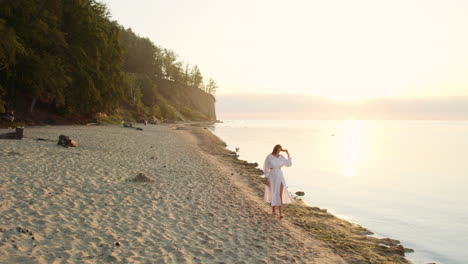 woman in white summer dress strolls by the sea at sunrise - aerial parallax slow motion