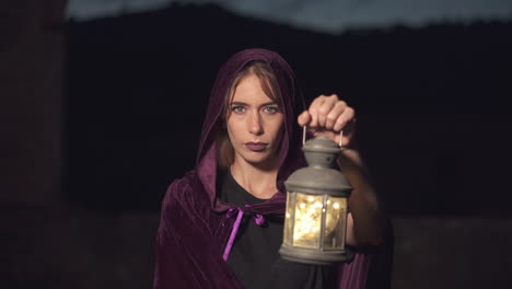 mysterious young woman in a purple cloak and a hood staring at the camera and using an old lantern at night