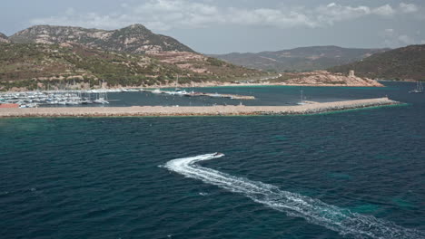 Ein-Boot-Rast-Durch-Das-Blaue-Wasser-In-Der-Nähe-Eines-Yachthafens-Mit-Hügeln-Im-Hintergrund-Auf-Sardinien