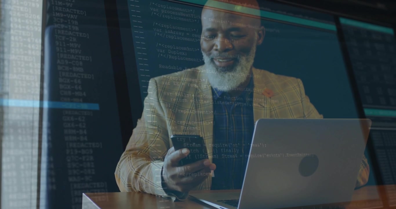 Senior tech professional man sitting at desk, holding smartphone viewing laptop with floating code