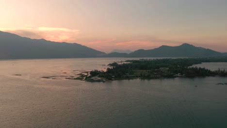 Puesta-De-Sol-Aérea-Volando-A-Través-Del-Pintoresco-Y-Pacífico-Paisaje-Asiático-Con-Lago,-Montañas-Y-Pueblo-De-Pescadores-En-Lang-Co,-Vietnam