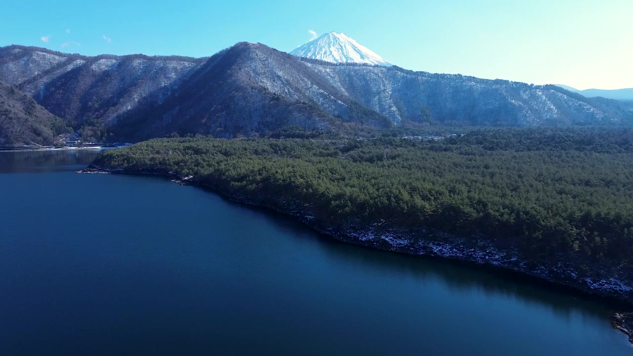 Ilmakuva dronella Saiko-järven vesiväylän joenlahdesta, jossa näkyy lumisia metsäisiä vuoria ja Fuji-vuoren huippu horisontissa Fujikawaguchikon Minamitsurun alueella Japanissa, luonnon helmassa.