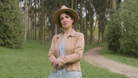 portrait of a middle aged woman in hat and crossed arms standing in the park and looking at the camera