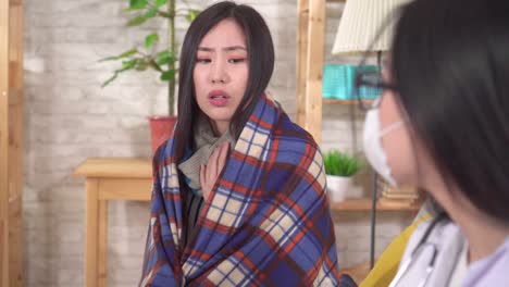 병든 아시아 여성은 플레이드와 장갑을 입고 열을 가지고 그녀의 거실에서 의사와 의사소통합니다.
