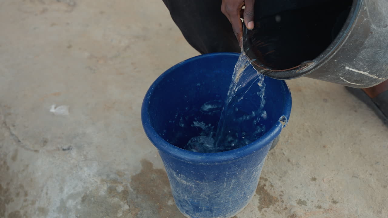 Trabalhador de tarefas domésticas derrama água de um balde preto para um balde azul em piso de concreto, com as mãos visíveis. Sequência repetida de derramamento, com pequenos salpicos e gotas, em limpeza de pátio externo