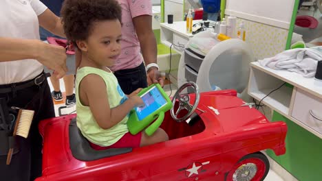 two year old adorable african european child having his first hair cut in a child hairdresser while playing with a ipad