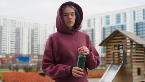 fitness enthusiast walking peacefully along pavement holding water bottle while looking around urban park setting with tall buildings and colorful spring flowers at corner on quiet tree lined path