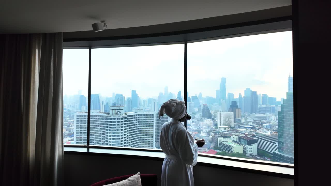 Woman in bathrobe enjoys Bangkok skyline view from high-rise hotel window