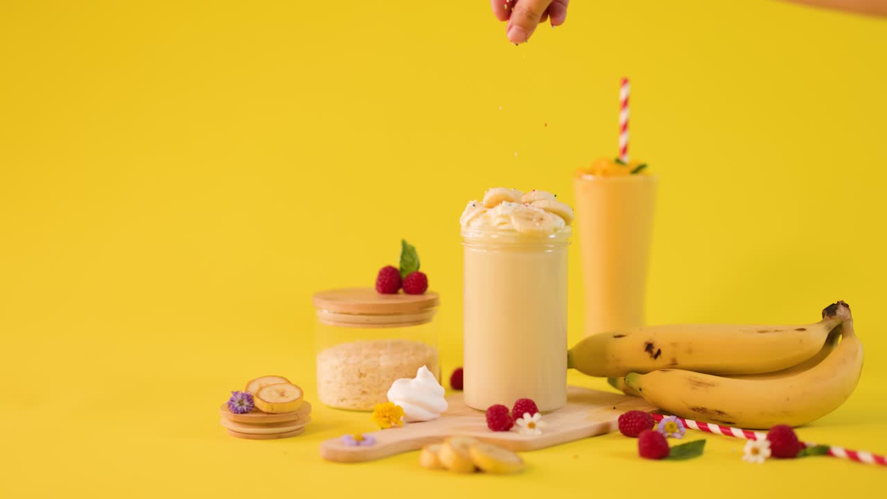 Una mano umana cosparge un frullato cremoso alla banana con guarnizioni e dessert alla frutta su uno sfondo giallo brillante