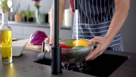 Midsection-of-biracial-man-wearing-apron-cleaning-vegetables-in-kitchen,-slow-motion