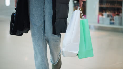 dama en pantalones vaqueros y chaqueta negra caminando en un espacio comercial bien iluminado llevando un bolso negro y dos bolsas de compras, fondo borroso con estantes y exhibiciones de tiendas en un centro comercial moderno