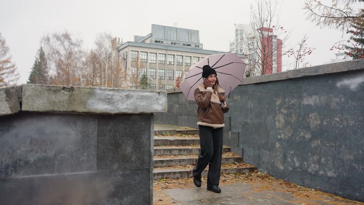 Happy girl smiling, making phone call while holding umbrella, wearing black knit cap, brown shearling jacket, black trousers, walking through urban park on cold day with light snowfall and autumn leaves