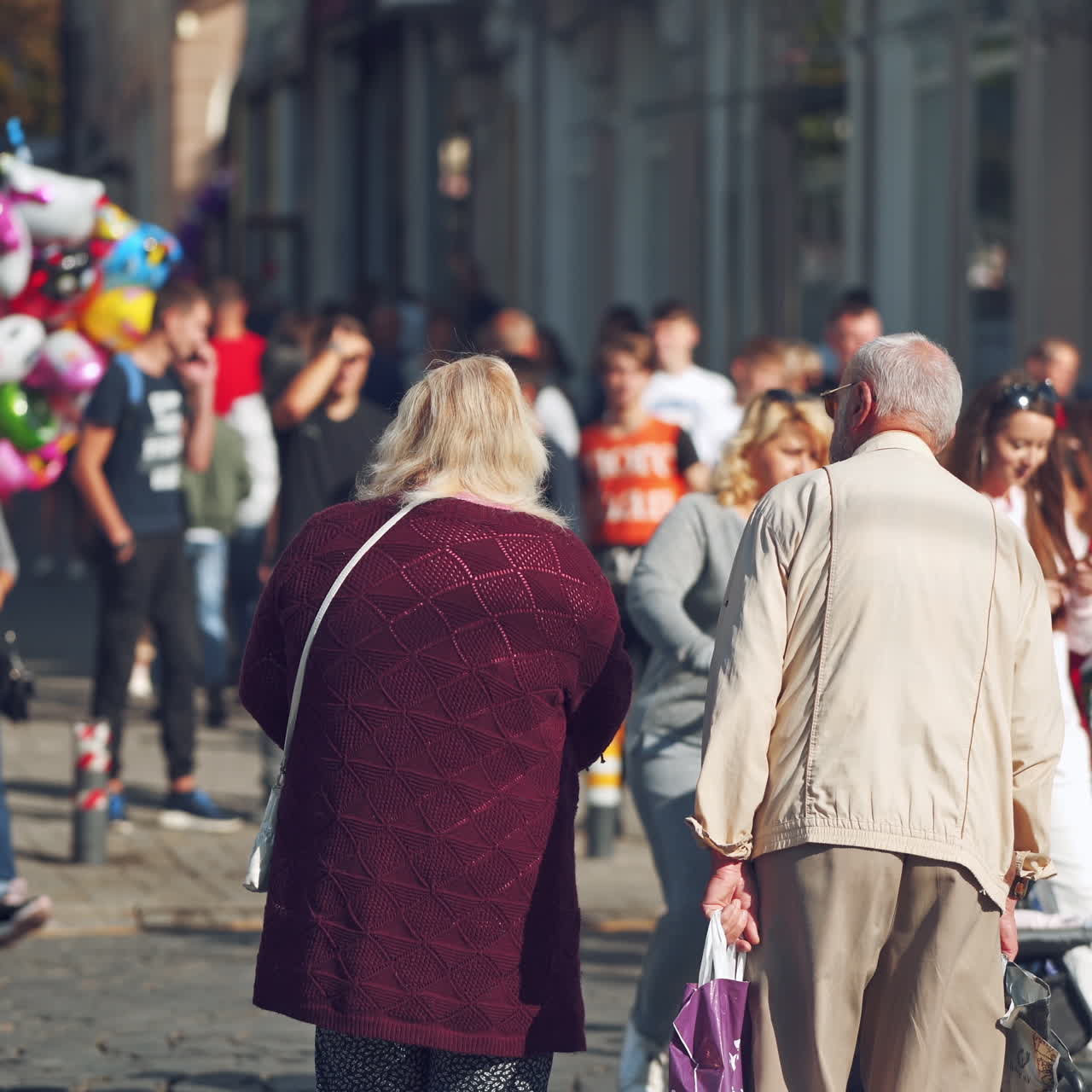 Many people in the city. Backside view of old people walking on the sidewalk on crowded city background. Colored balloons sold in the street.
