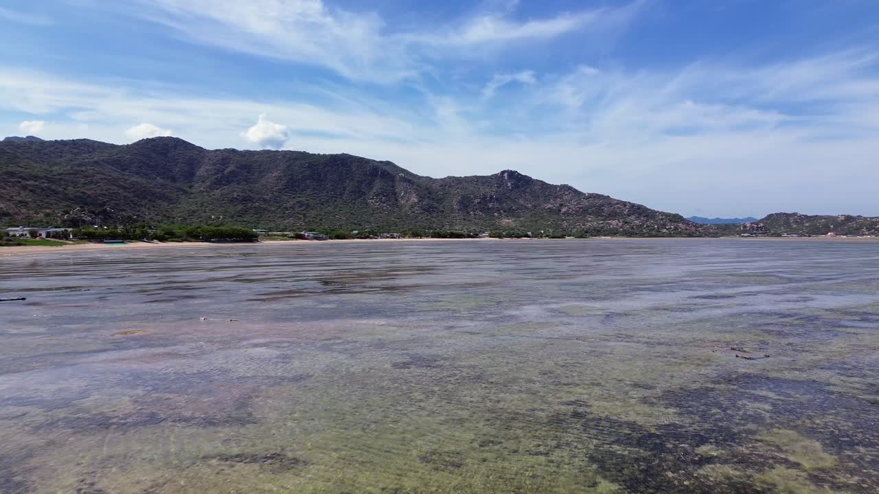 Drone advances and pans right simultaneously, showcasing the serene My Hoa Lagoon with mountains and beach in the background.