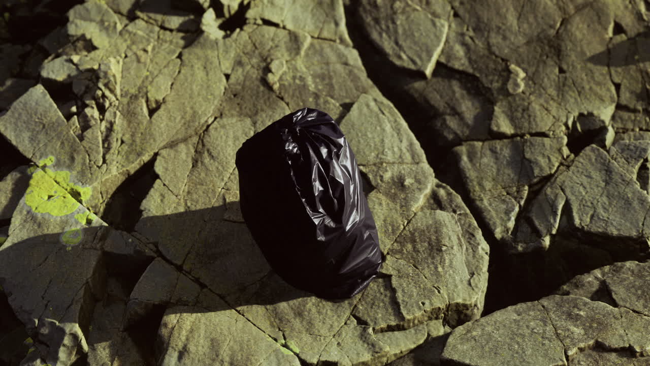 Black trash bag rests on rugged stones under the afternoon sun by the riverbank