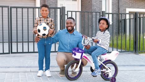 retrato de feliz padre afroamericano sonriendo a la cámara con niños pequeños en la calle en las afueras