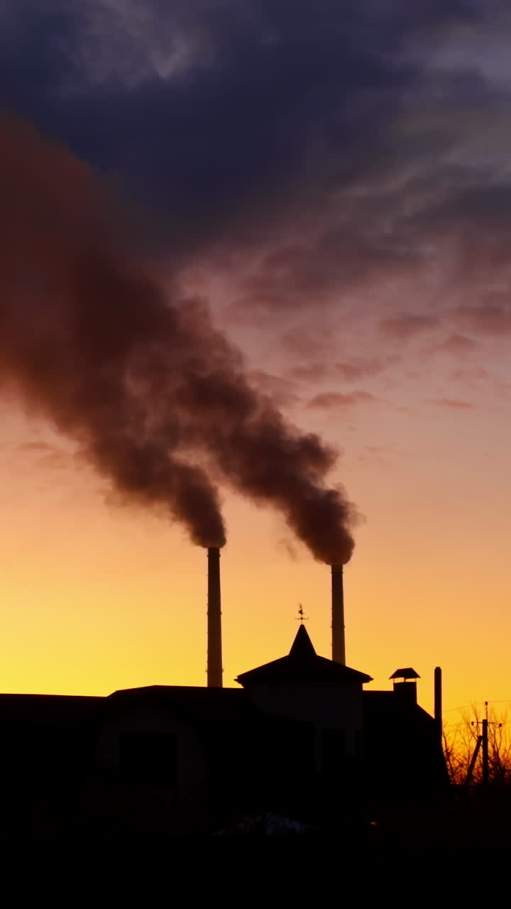 Two pipes rising above the building. Heavy dark smoke gathering in the atmosphere. Sunset landscape. Vertical video