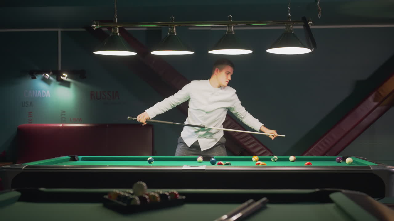Giovane uomo in camicia bianca e pantaloni grigi si sporge su un tavolo da biliardo verde, impugnando la stecca con intensa concentrazione mentre allinea il tiro. Le luci dall'alto proiettano un bagliore sull'elegante sala da biliardo con sedute rosse