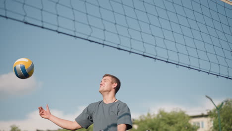 young man with plaid cloth around waist jumps to hit volleyball but misses, smiling in response, bright outdoor court with trees and building in background