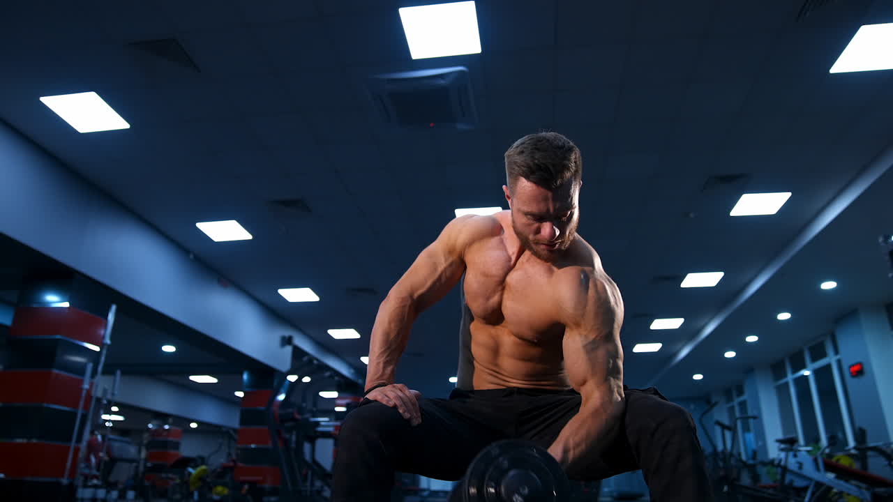 Handsome power athletic man bodybuilder doing exercises with dumbbells. Fitness muscular body on dark gym background. Slow motion.