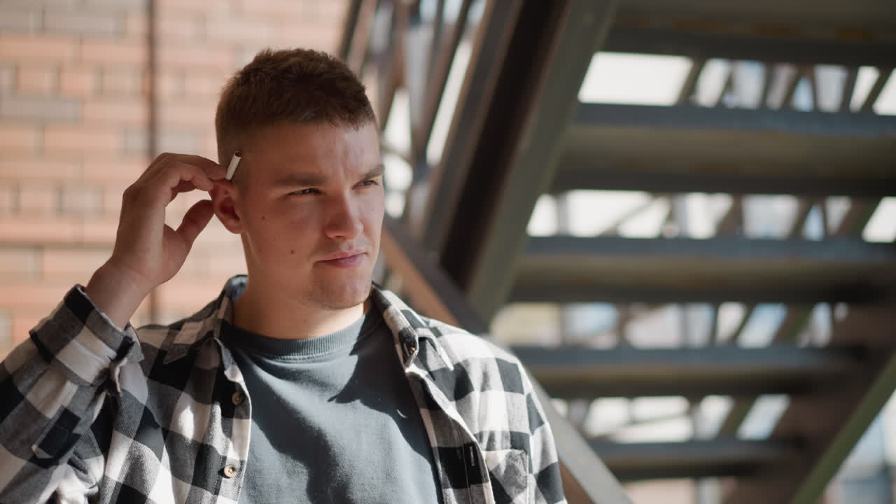 portrait view of student in black white plaid shirt on metal staircase returning cigarette to ear while looking at camera against sunlit brick wall and blurred staircase under midday sun