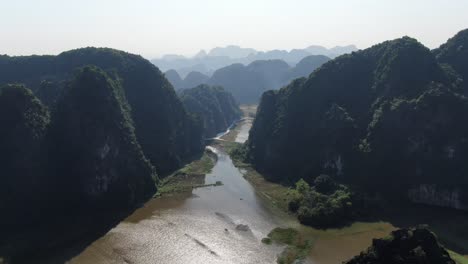 drone aerial view in vietnam flying through rocky mountains covered with green trees over a wide brown river in ninh binh on a sunny day