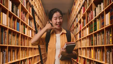asian woman student with a backpack and some books smiling and showing call me gesture while standing in the library