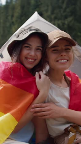 two women embrace with a rainbow flag at a campsite