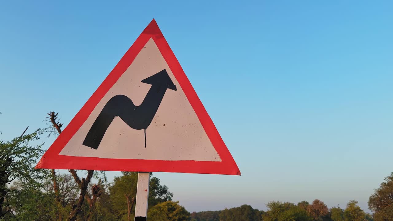 Inquadratura ravvicinata orbitante su un segnale di pericolo triangolare di strada tortuosa montato su un palo a bordo strada, con un cielo azzurro limpido e delicati elementi di sfondo rurali