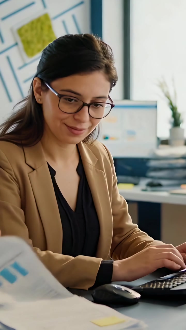 Video vertical: Mujer tecleando con blazer color canela revisando gráficos de un colega mientras escribe en el teclado.