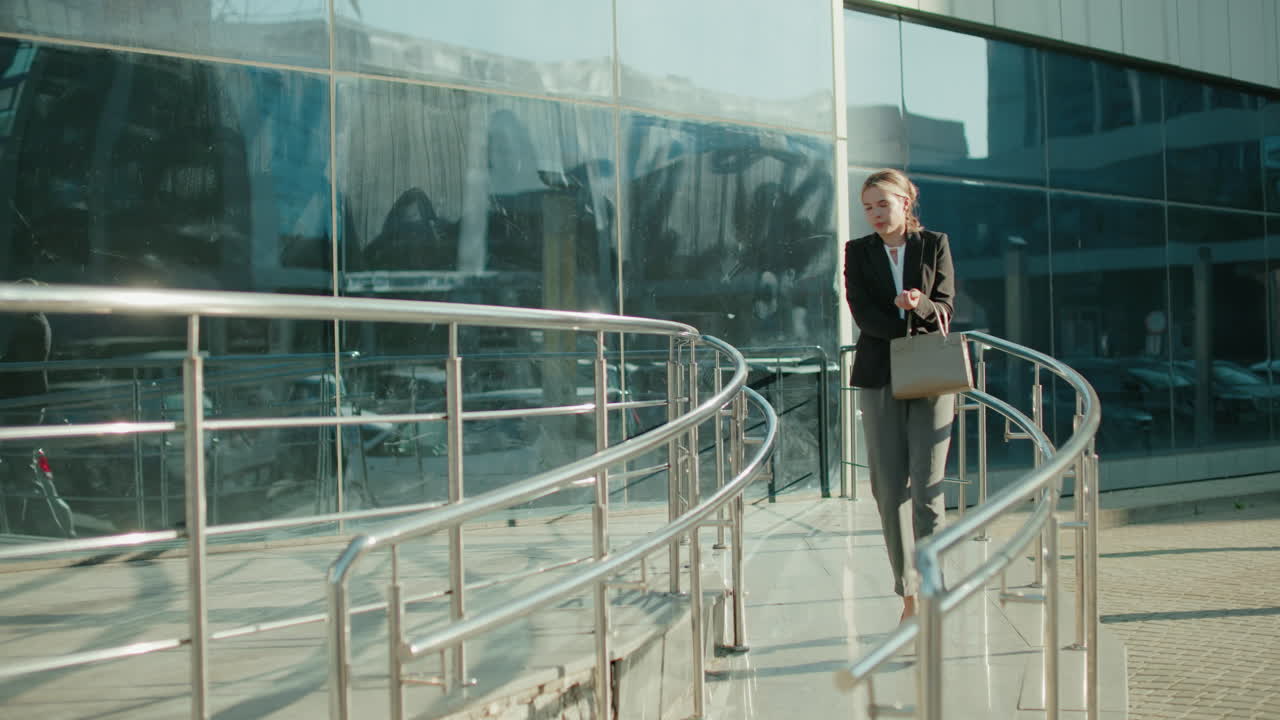 Young woman walking down office ramp after work, contemplatively checking handbag to get car key, with glass building reflecting surroundings, bright sunlight casting shadows on pavement