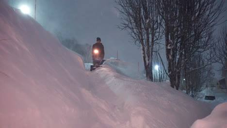 man using a blue snowblower to clear deep snow at night. bright artificial light illuminates snowy path. static view