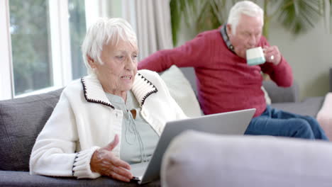 happy senior caucasian couple sitting on sofa using laptop for video call at home, slow motion