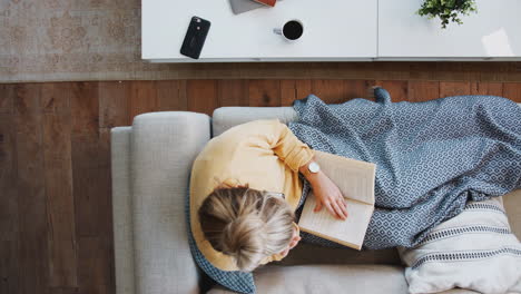 overhead shot looking down on woman at home lying on reading book and drinking coffee