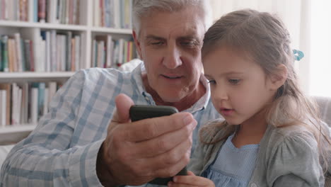 Glücklicher-Großvater-Zeigt-Kleinem-Mädchen,-Wie-Man-Das-Smartphone-Benutzt,-Und-Lehrt-Neugierige-Enkelin-Moderne-Technologie.-Intelligentes-Kind-Lernt-Mobiltelefon,-Das-Mit-Opa-Auf-Dem-Sofa-Sitzt